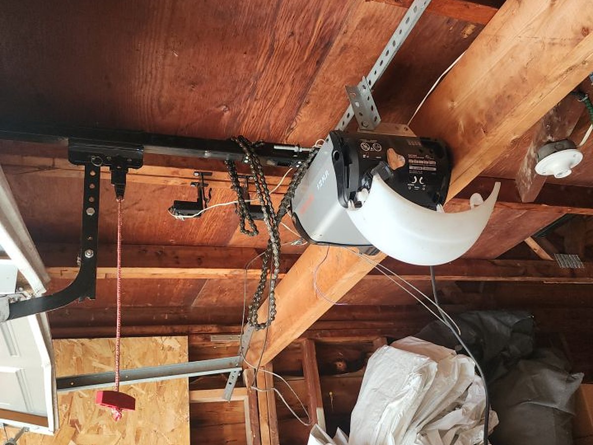 Chain-drive opener bolted to the exposed rafters of an unfinished garage ceiling, with the rail, chain and manual release cord in view
