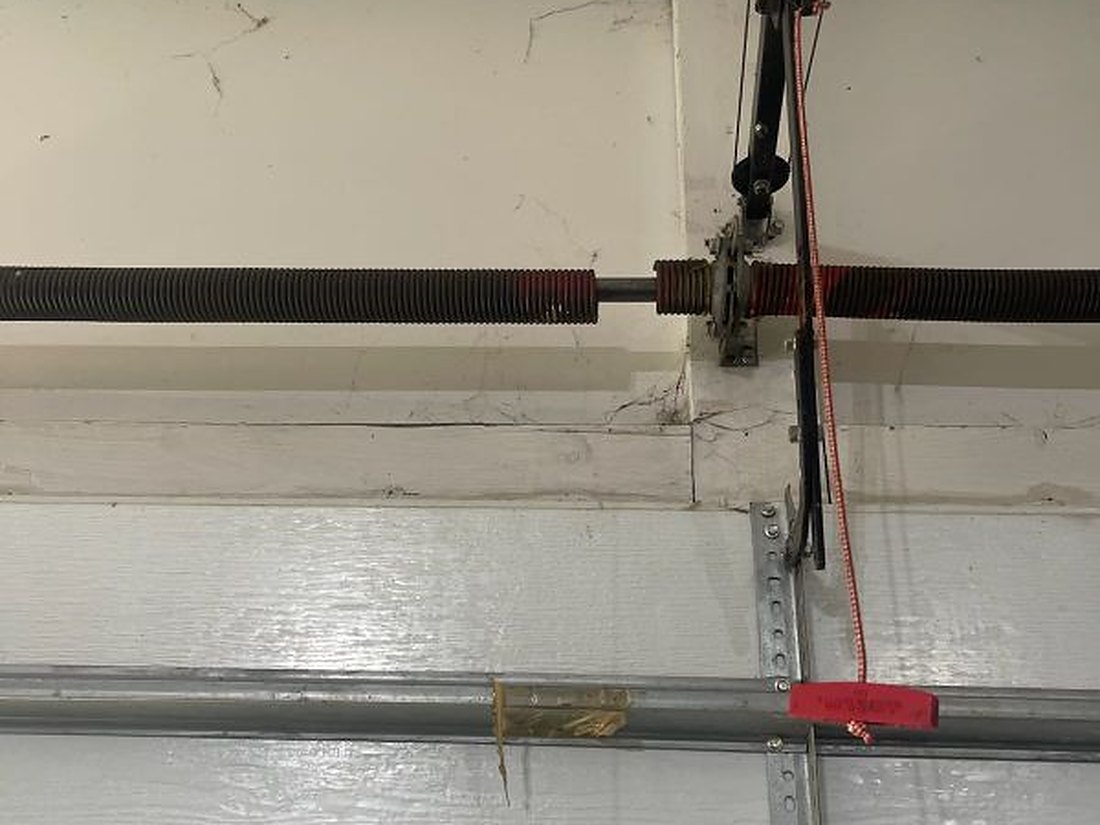 Torsion springs and shaft mounted across the header above a closed residential garage door, with the cable drums and the red manual release cord in view