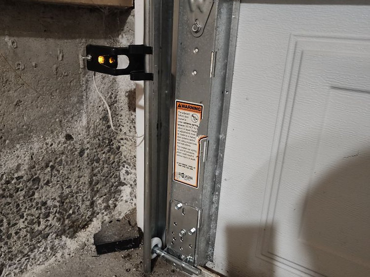 Bottom of a vertical garage door track with the roller bracket and lift cable, and the wall-mounted safety sensor beside the opening